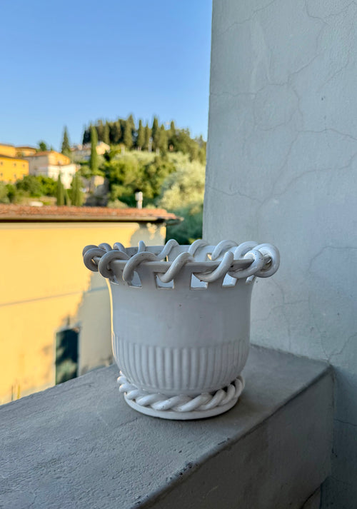 A white ceramic planter with a braided rope-style rim and matching base, placed on a concrete ledge outdoors with a blurred background of buildings, trees, and blue sky.