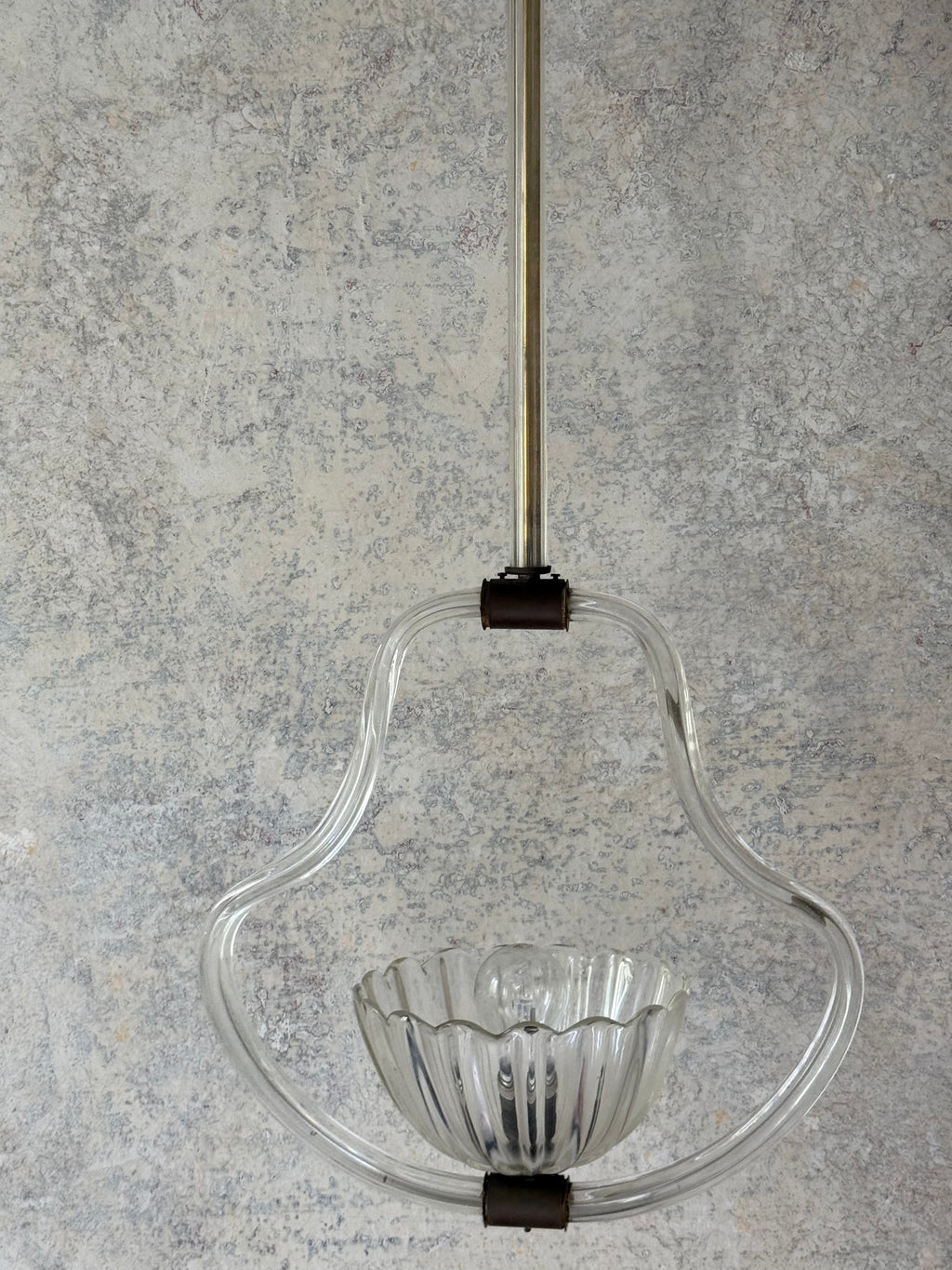 A clear glass pendant light fixture with a ribbed glass bowl and a curved glass frame, suspended by a polished metal rod with dark metal connectors against a textured gray plaster wall.