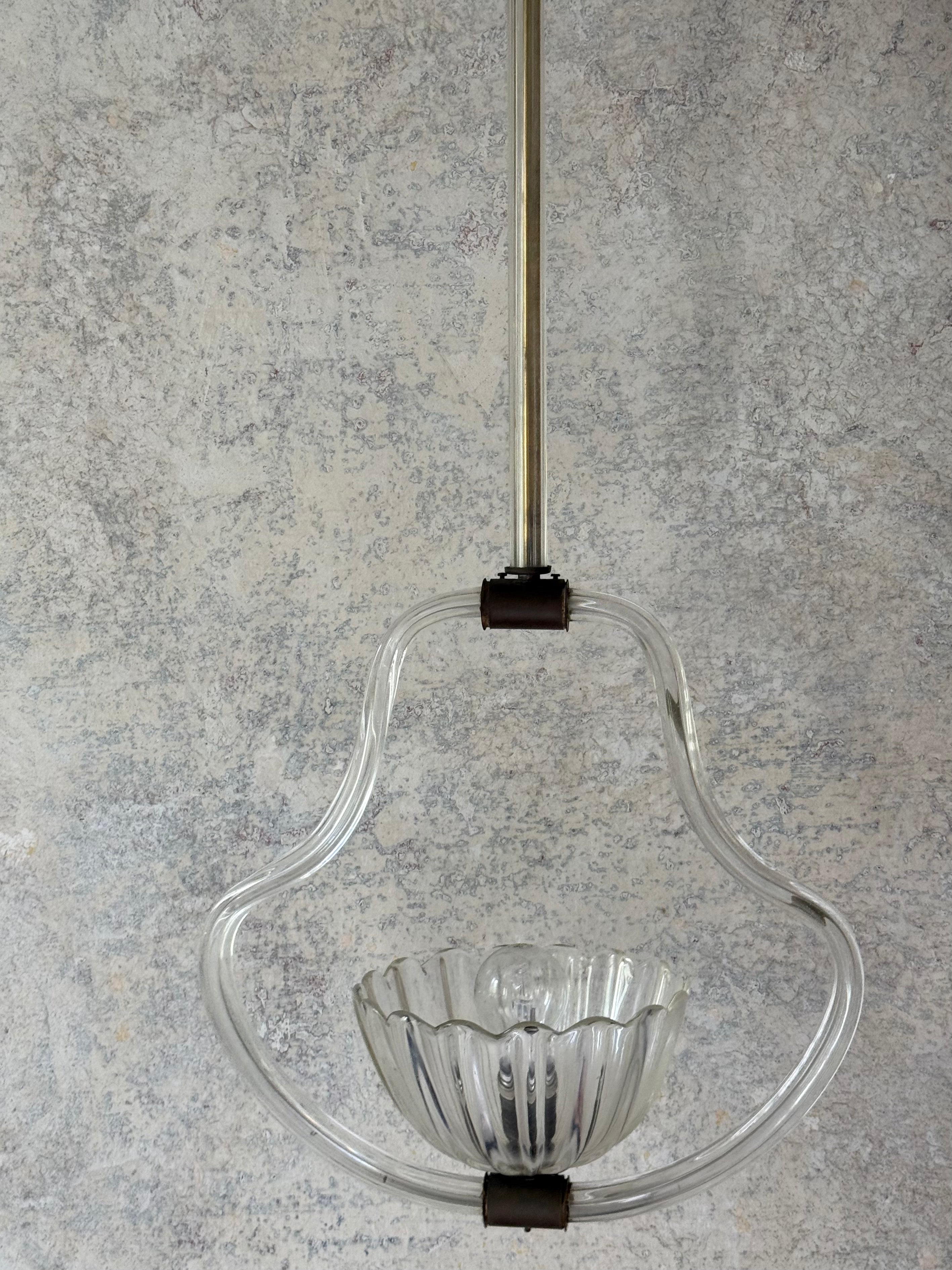 A clear glass pendant light fixture with a ribbed glass bowl and a curved glass frame, suspended by a polished metal rod with dark metal connectors against a textured gray plaster wall.