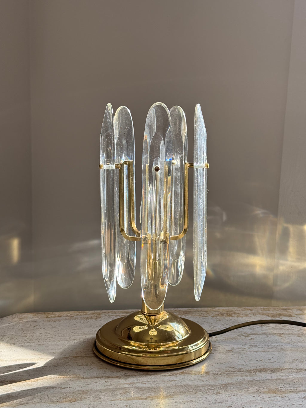 A mid-century modern table lamp featuring a polished gold brass base and multiple clear glass or acrylic prisms, set on a light stone surface against a neutral background.