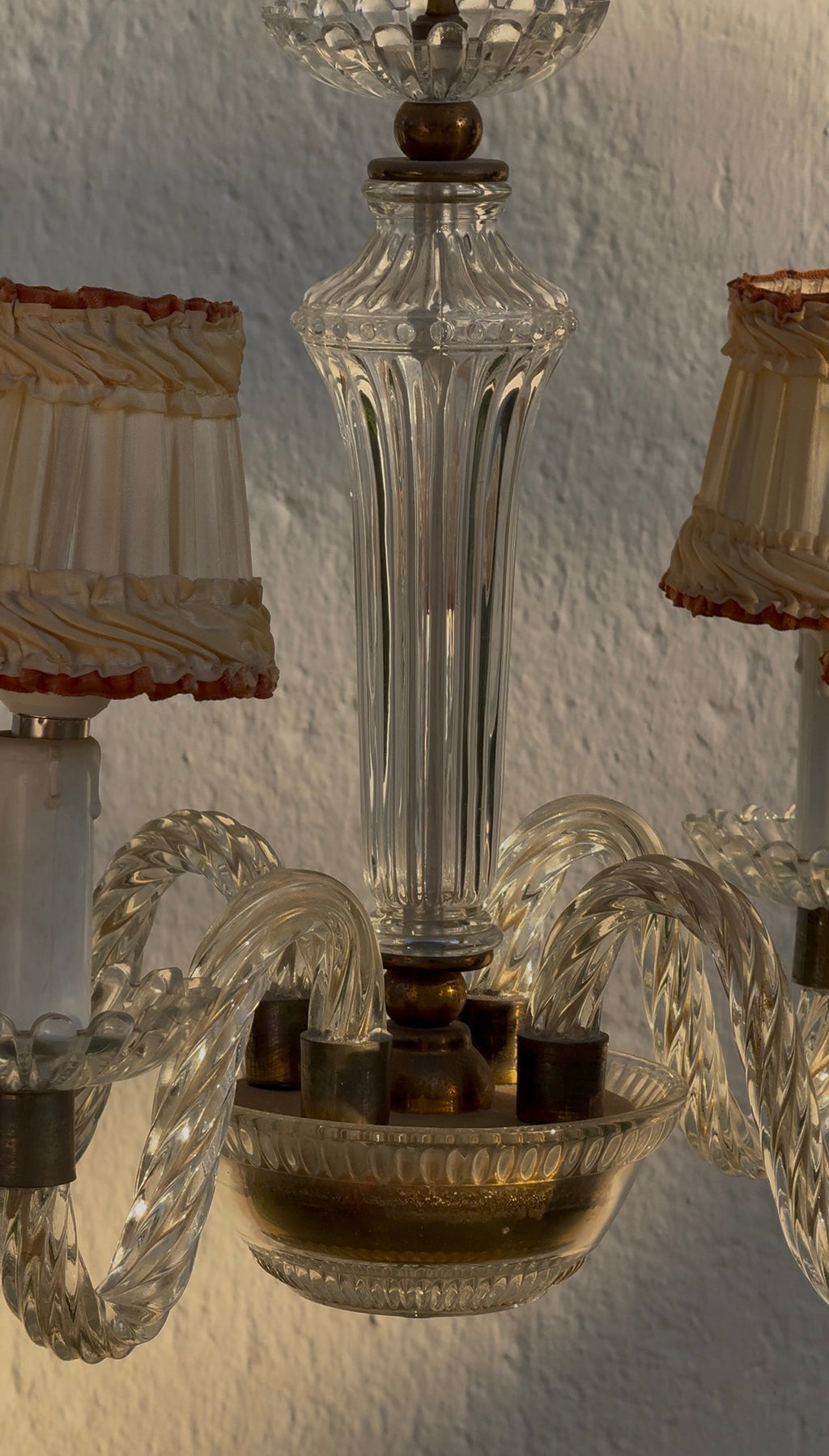 A close-up of a clear glass chandelier with twisted glass arms and a fluted glass stem, shown against a textured white wall with warm light.