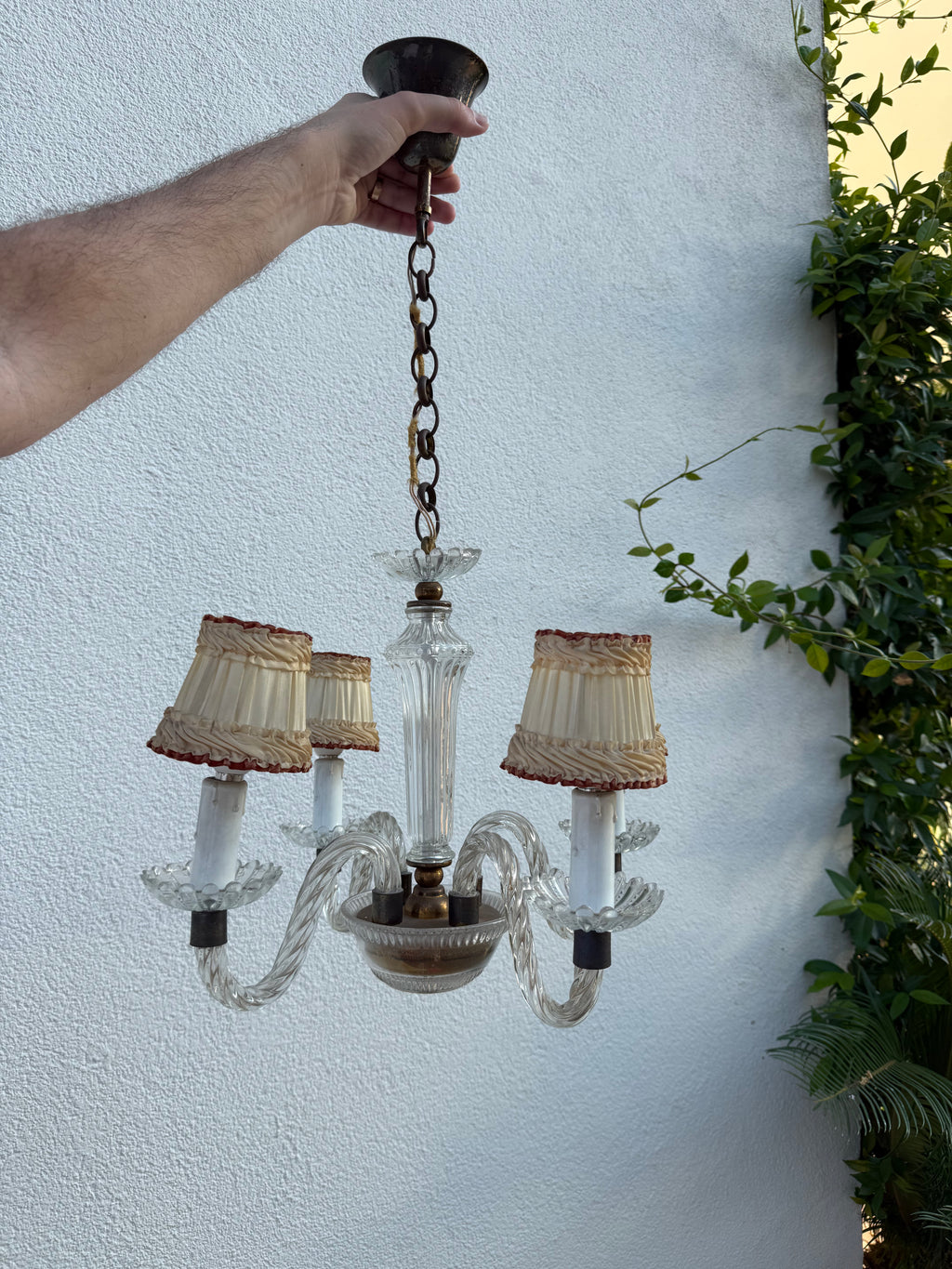 A three-light vintage-style chandelier with clear glass arms and shades, held by a person's hand against a white stucco wall with green plants on the right.
