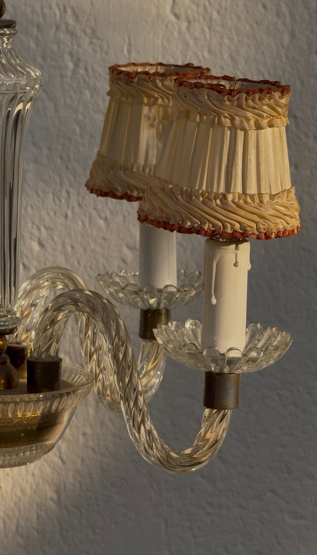 A close-up of a vintage-style wall sconce with two cream pleated fabric shades trimmed in brown, mounted on a clear glass curved arm with white candle-style sockets.