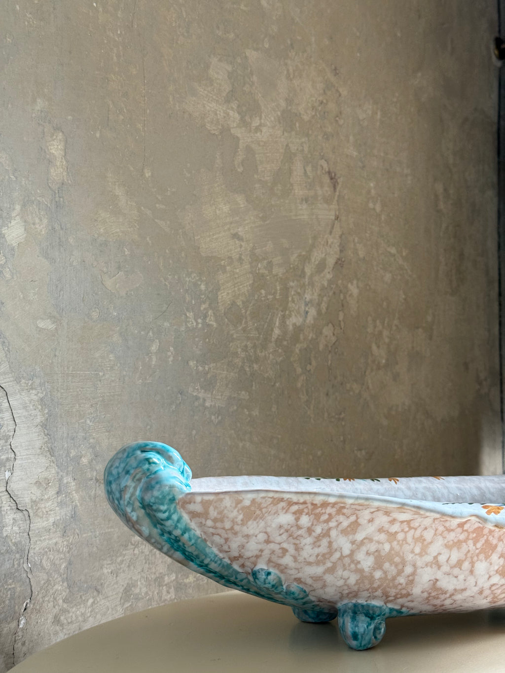 A decorative ceramic bowl with a white and peach marbled glaze and turquoise blue raised edges, shown on a tabletop against a textured beige wall.