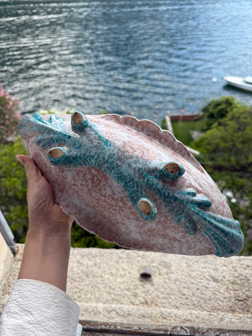 A hand holds a decorative ceramic bowl shaped like a stylized lizard, featuring a pink speckled glaze and turquoise blue accents, with a scenic outdoor background of water and greenery.
