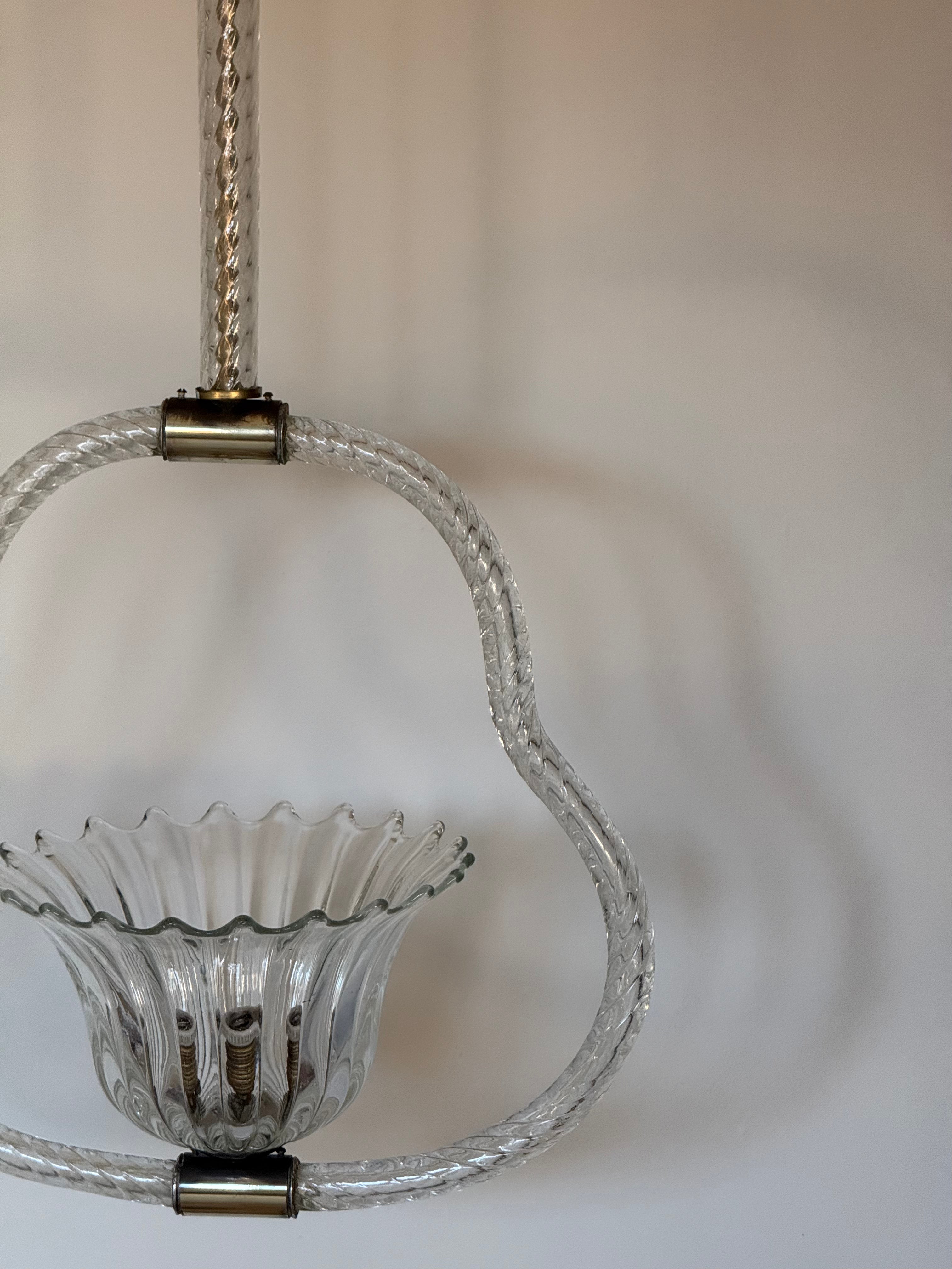 A close-up of a hanging glass chandelier or pendant light with a clear ribbed glass bowl, a curved clear glass loop, and brass-toned metal hardware against a plain light background.
