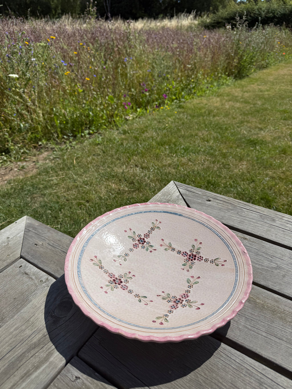 A round ceramic serving platter with a scalloped pink rim and floral patterns, resting on a weathered wooden table in an outdoor garden setting.