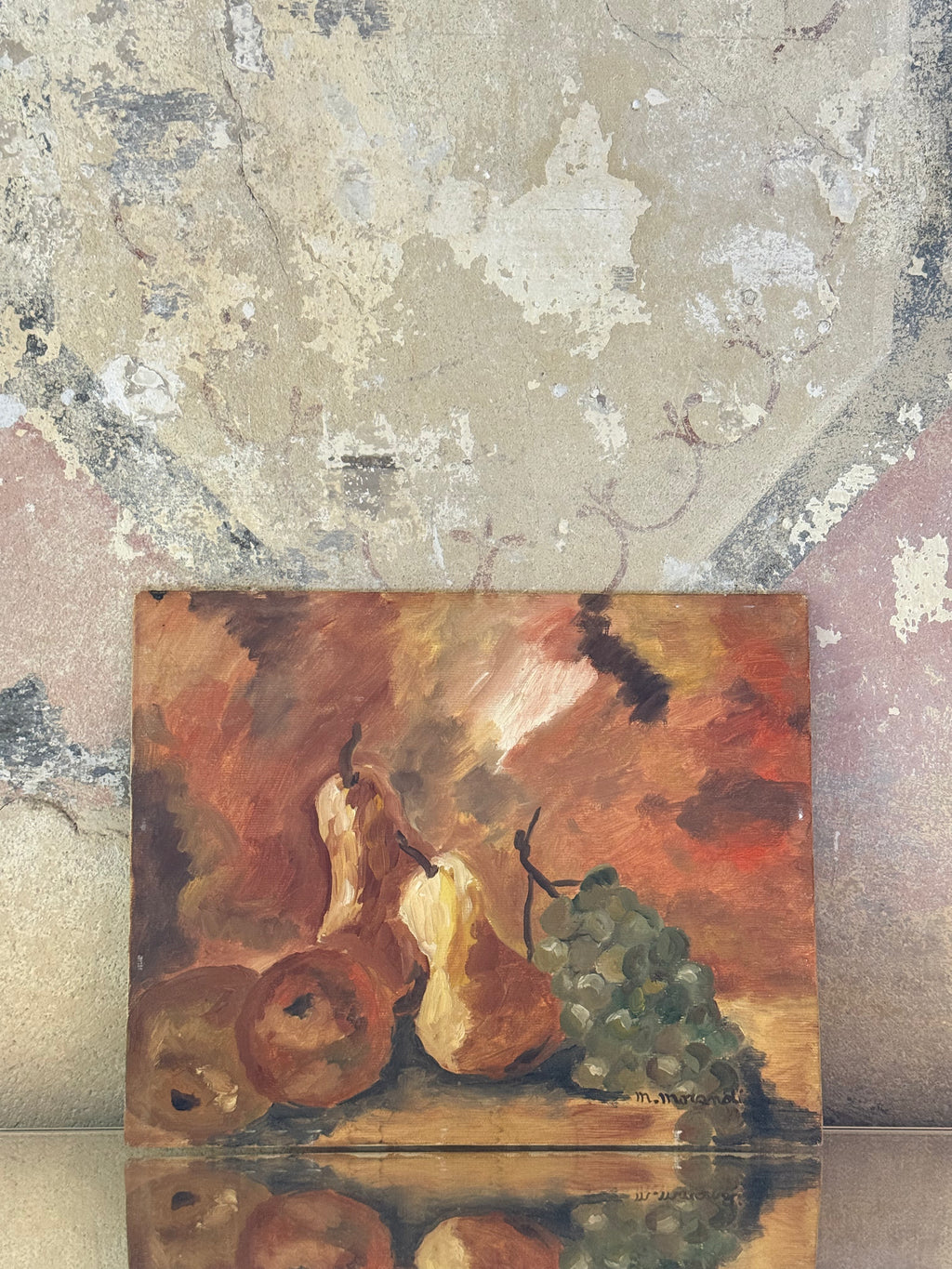 A square oil painting depicting a still life of pears, apples, and a bunch of grapes, displayed on a reflective surface against a textured, distressed plaster wall.