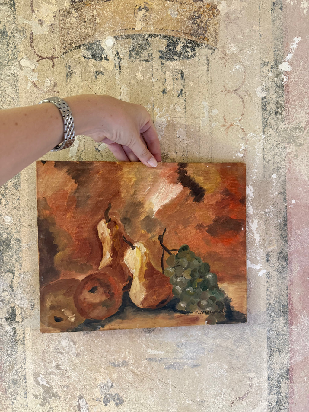 A hand holds a small still life oil painting depicting pears, apples, and a bunch of grapes, set against a textured, distressed beige background.