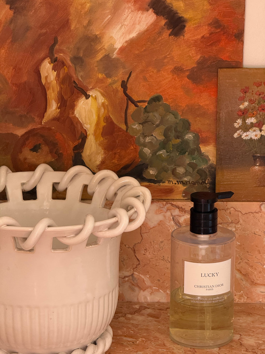 A bottle of Christian Dior Lucky hand wash with a black pump and clear cap, set on a terracotta countertop next to a white ceramic planter in front of a warm-toned still life painting.