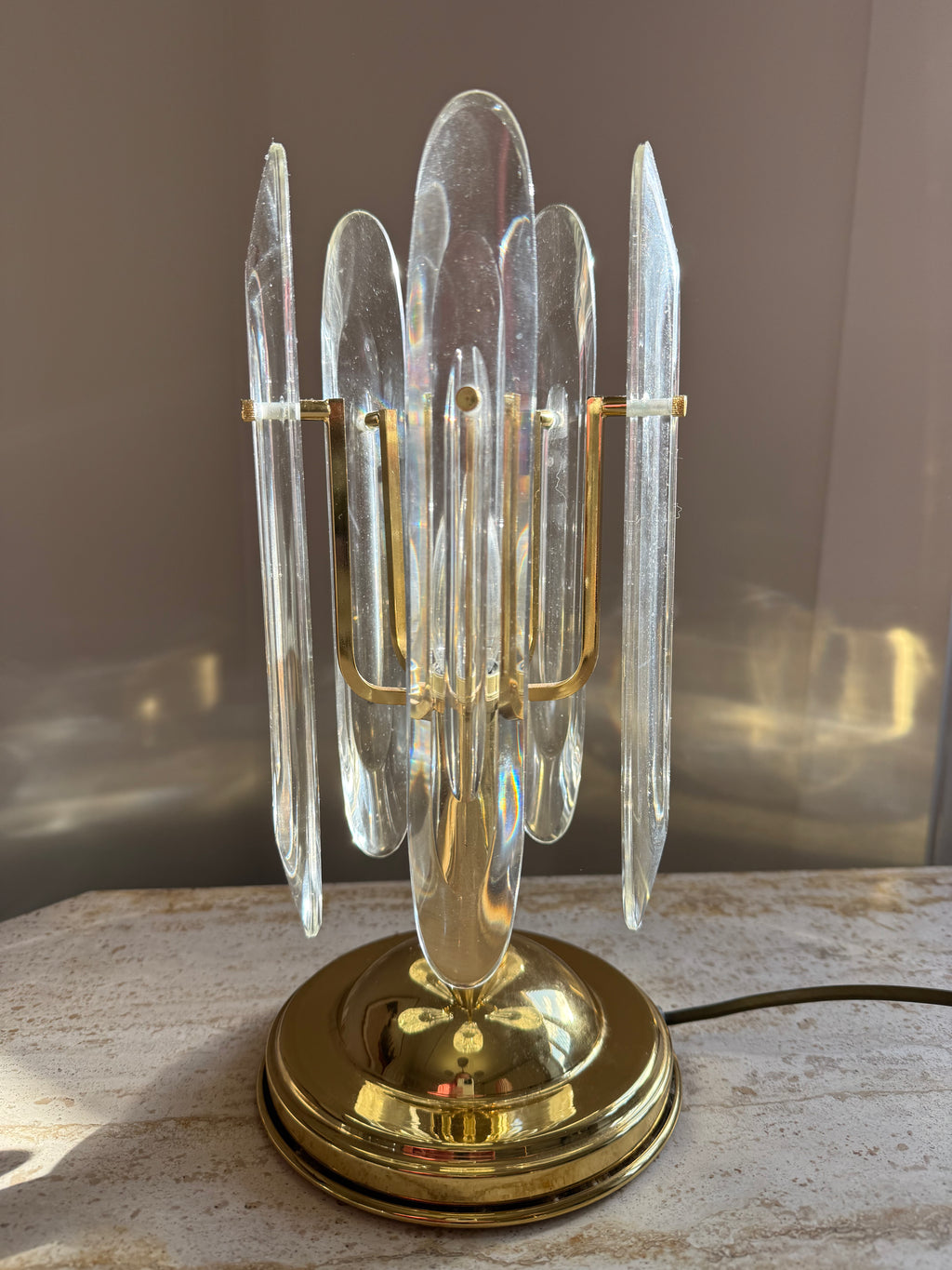 A brass table lamp with multiple clear glass or acrylic crystal-like vertical prisms arranged in a cluster, sitting on a light stone surface against a neutral indoor background.