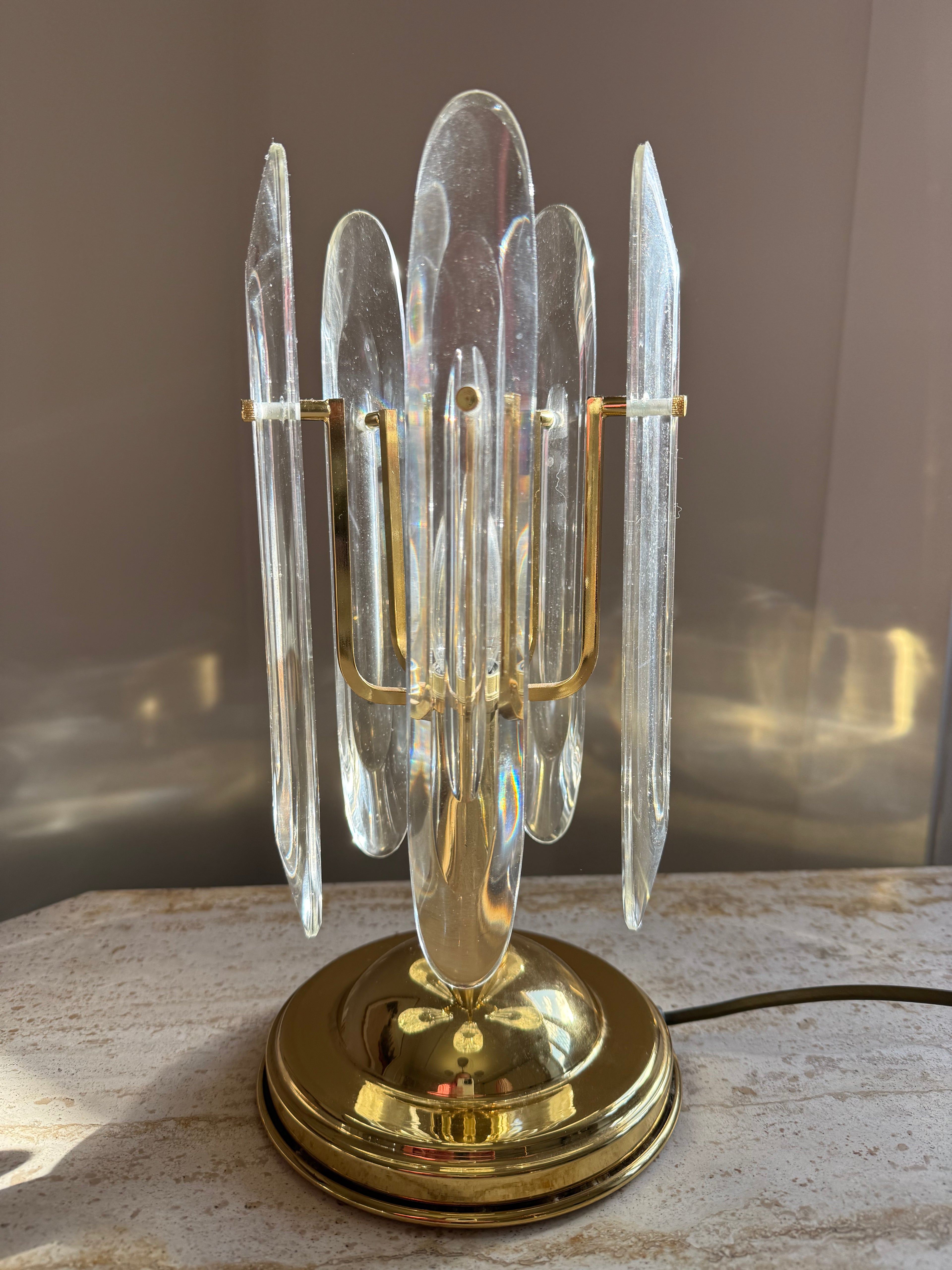 A brass table lamp with multiple clear glass or acrylic crystal-like vertical prisms arranged in a cluster, sitting on a light stone surface against a neutral indoor background.