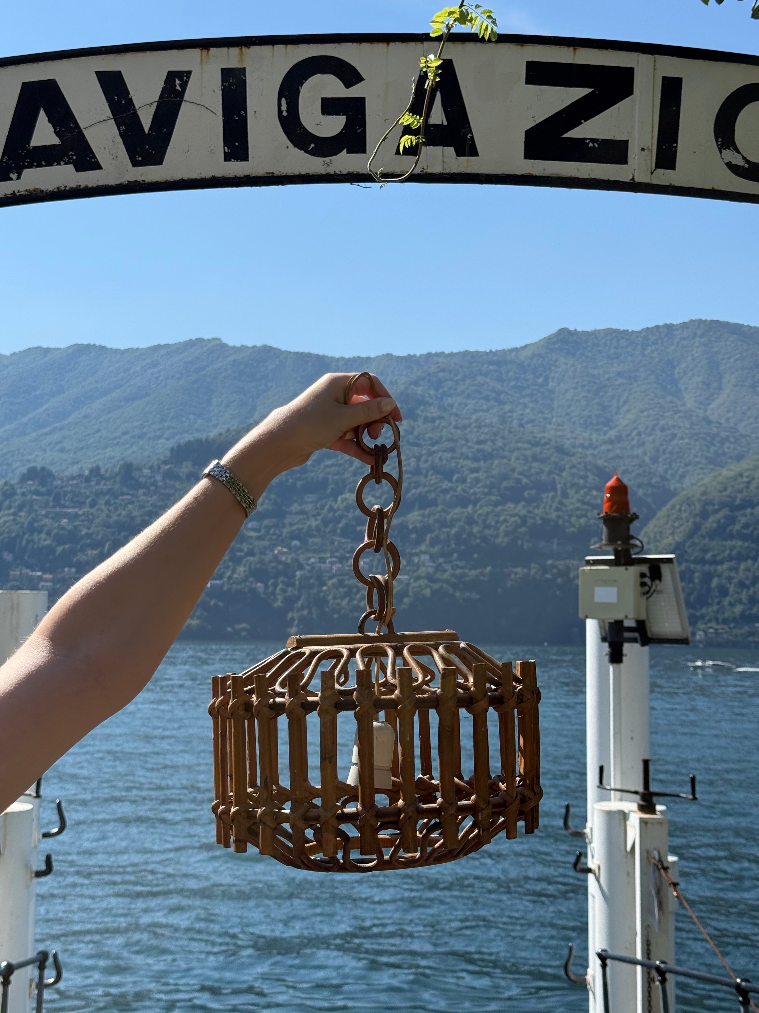 A hand holds a round wooden hanging lantern or cage-style light fixture outdoors by a lake, with a large sign reading NAVIGAZIO above and mountains in the background.