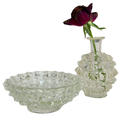 A clear textured glass bowl and matching small glass vase with a dark burgundy rose stem, photographed on a plain white background.
