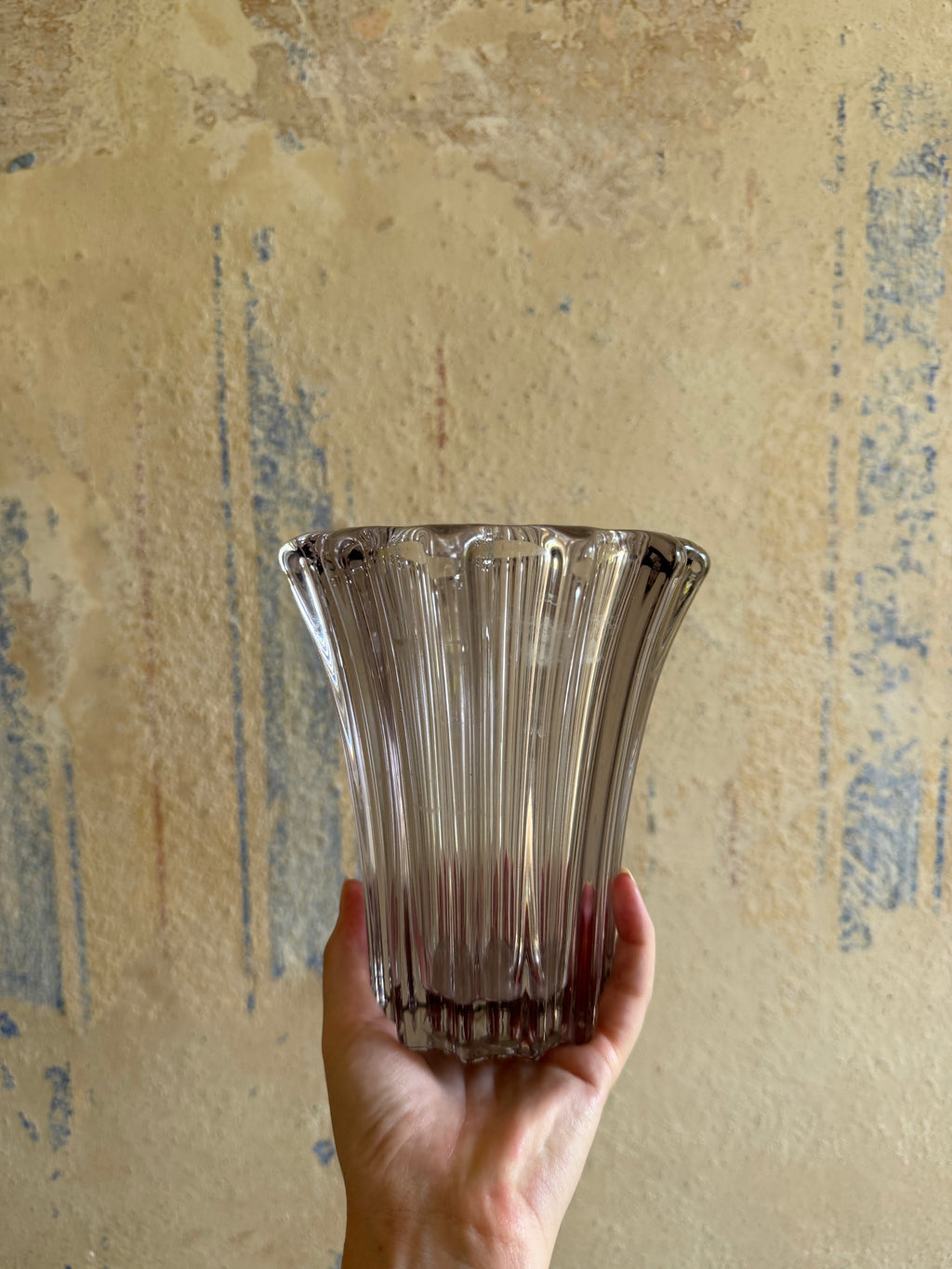A hand holding a ribbed, flared clear glass vase against a textured beige wall with blue distressed patches.