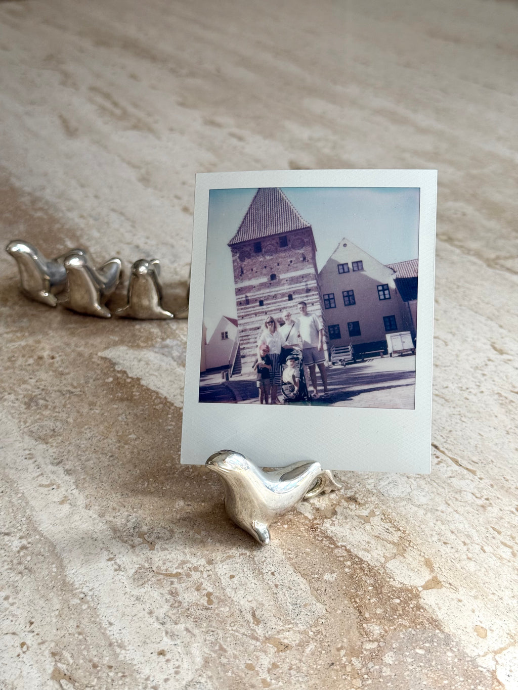 A square instant photo print with a white border stands upright on a small silver bird-shaped holder on a textured beige stone surface. The printed photo shows an outdoor scene with a tower building and two people standing in front of it.