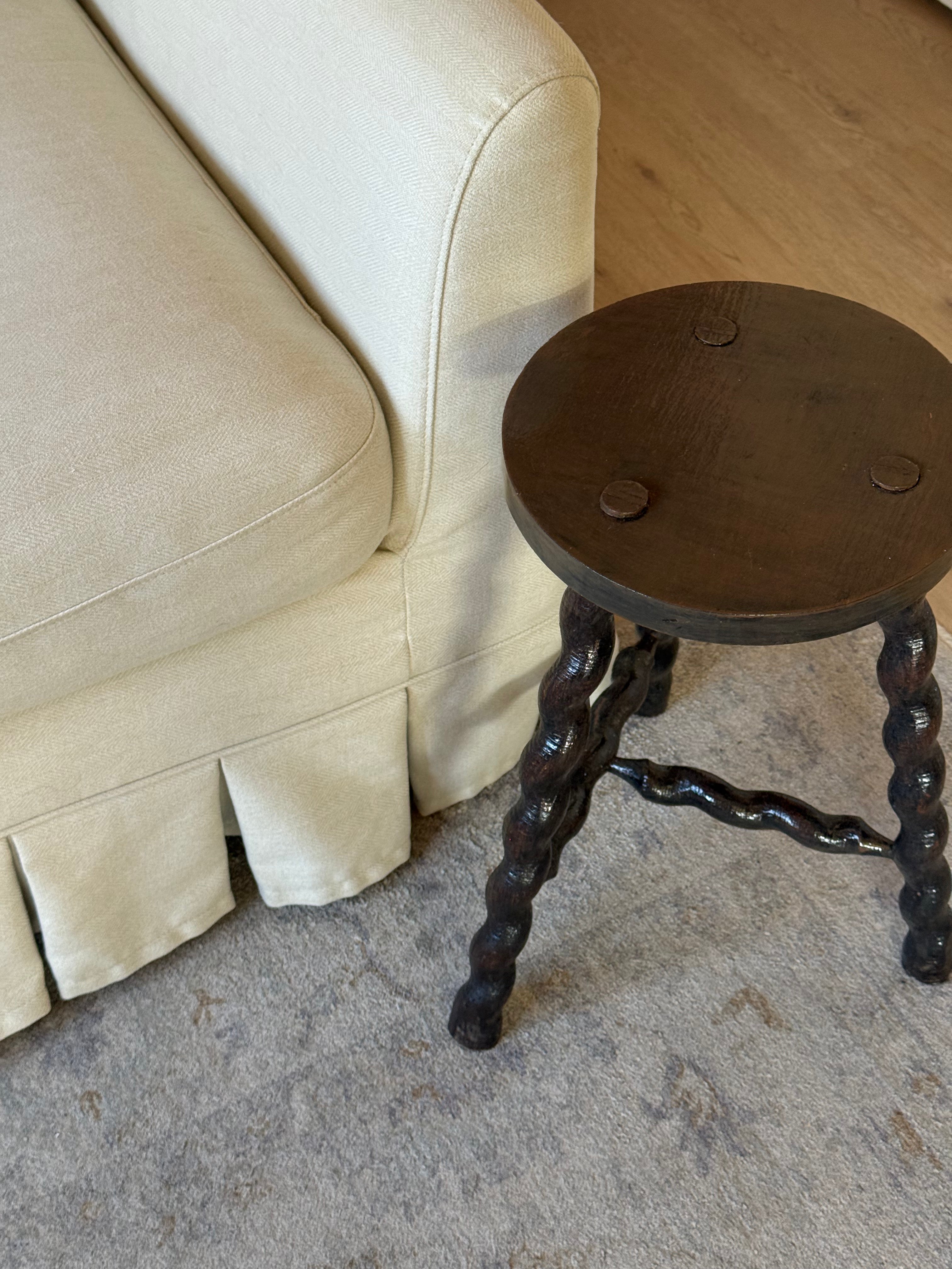A dark brown wooden stool with twisted legs and three metal cup holders on the seat, positioned next to a cream-colored slipcovered sofa on a gray carpeted floor.