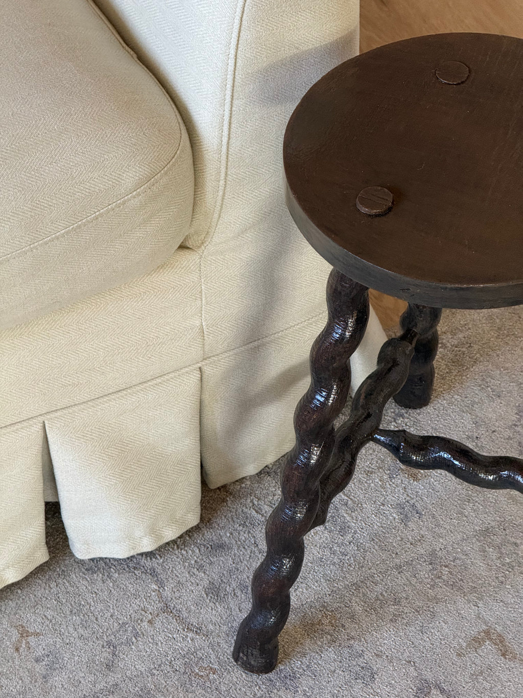 A close-up of a rustic dark wood side table with twisted, gnarled legs placed next to a cream-colored slipcovered sofa on a textured rug.