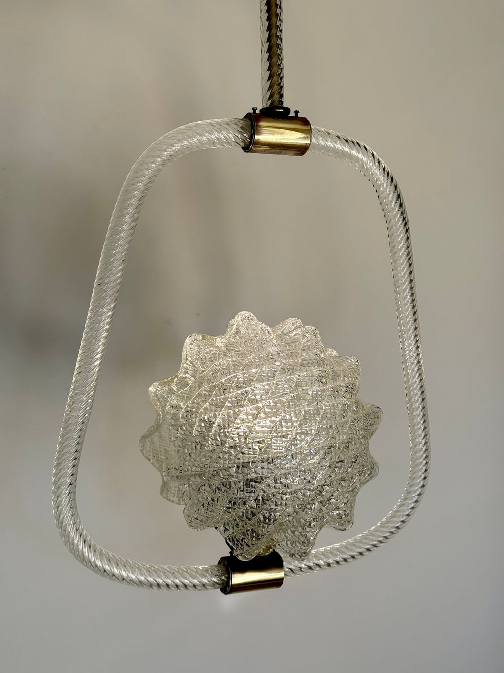 A hanging pendant light with a textured clear glass shell at the center, enclosed in a twisted clear glass frame, suspended by a black cord with brass-colored metal caps against a neutral gray background.