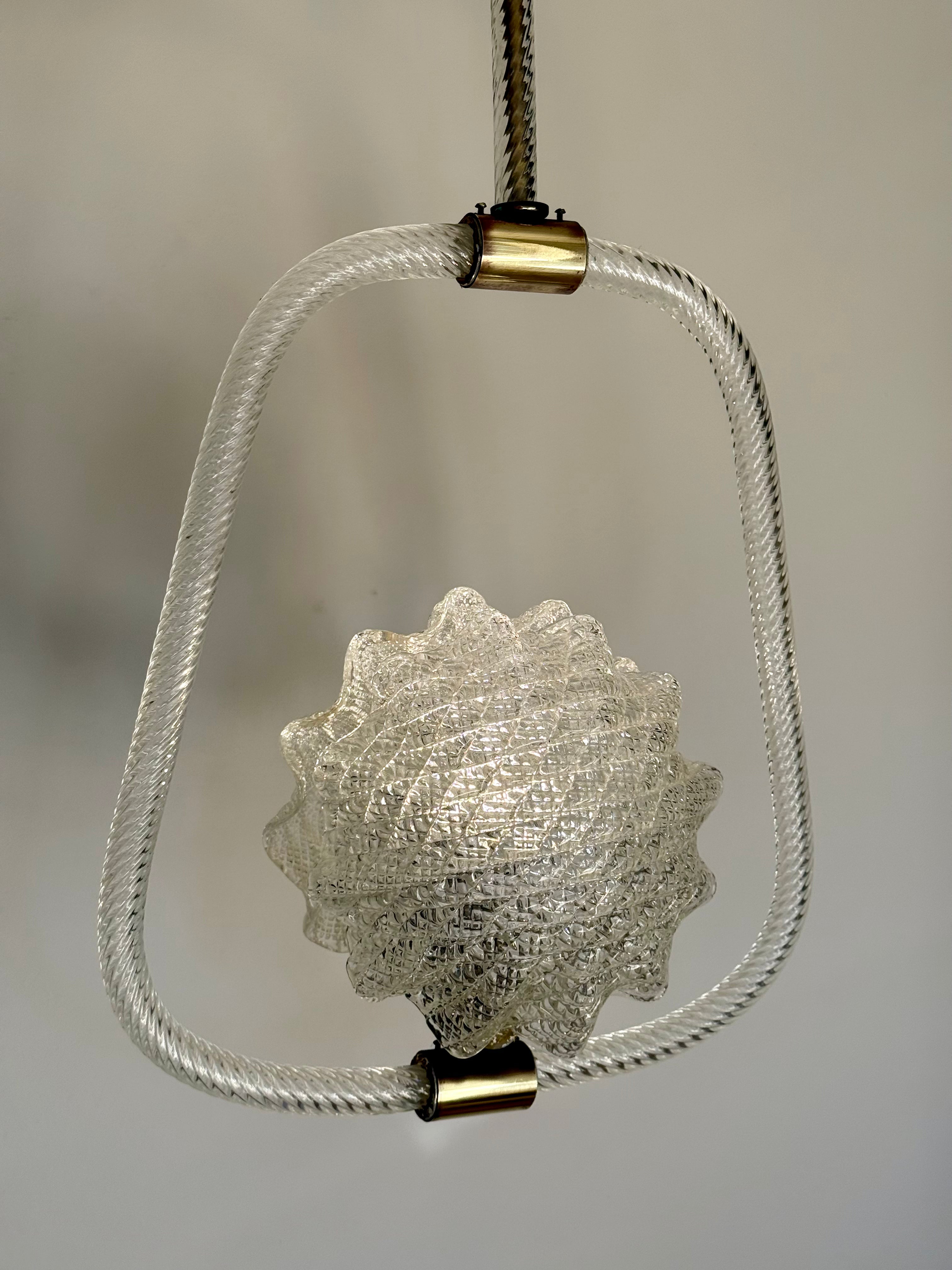 A hanging pendant light with a textured clear glass shell at the center, enclosed in a twisted clear glass frame, suspended by a black cord with brass-colored metal caps against a neutral gray background.