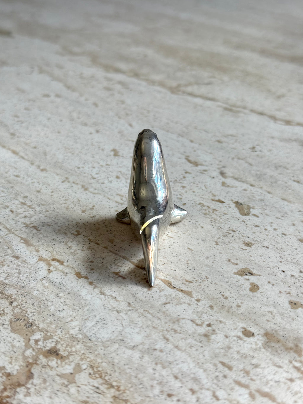 A small polished silver dolphin figurine standing upright on a light beige stone surface.
