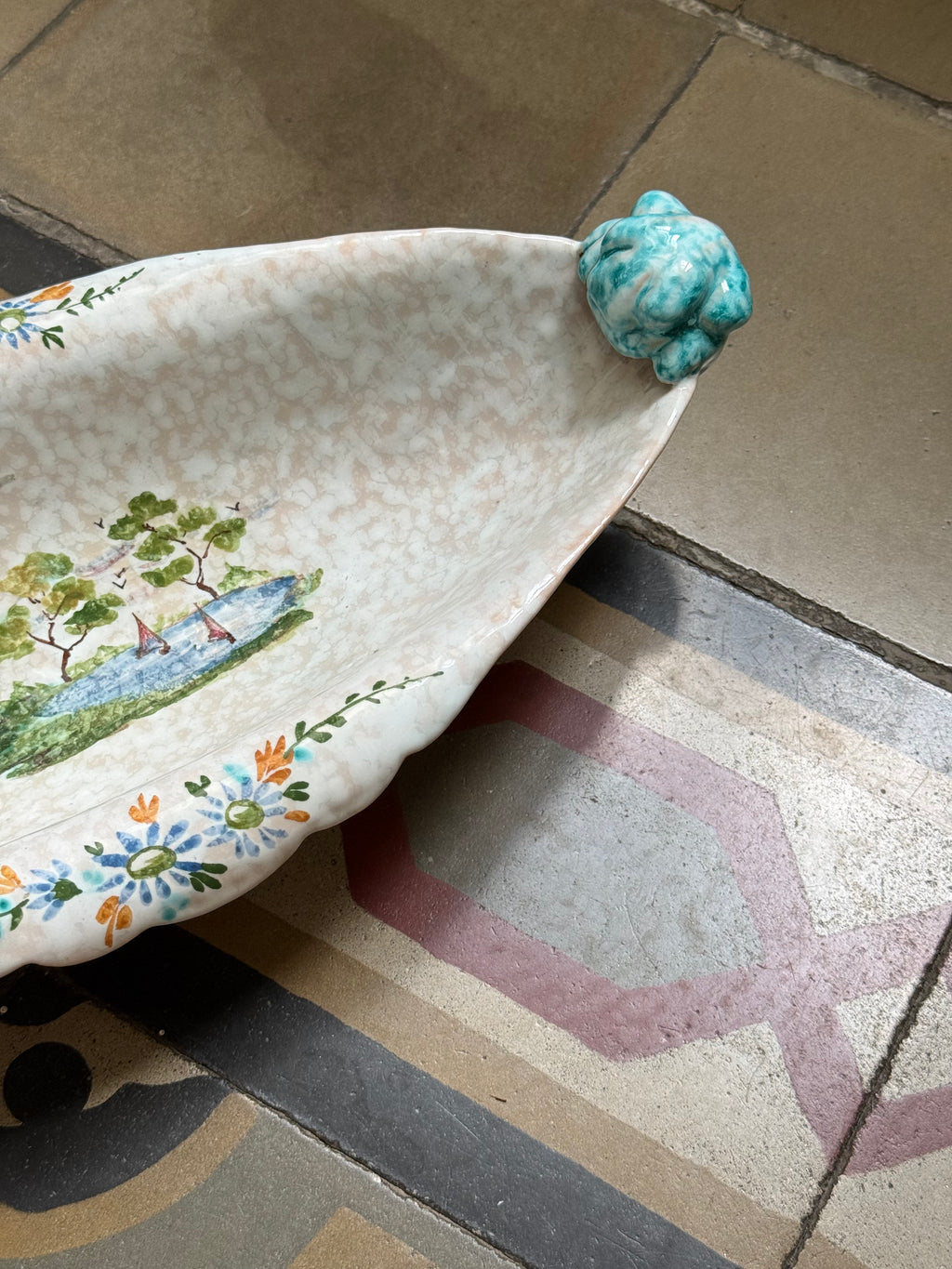 A ceramic leaf-shaped serving bowl with a hand-painted landscape and floral border, featuring a small turquoise frog figurine on the rim, set on a patterned tile floor.