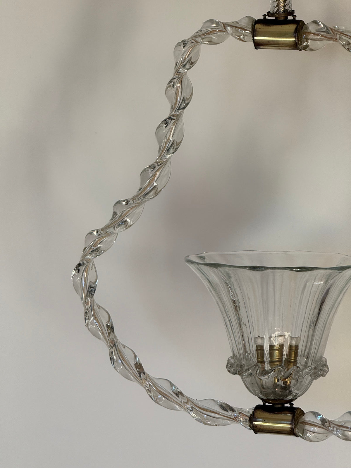 Close-up of a clear glass chandelier arm with a twisted, spiral-shaped glass rod and a flared ribbed glass cup shade, accented with brass metal caps against a neutral light gray background.