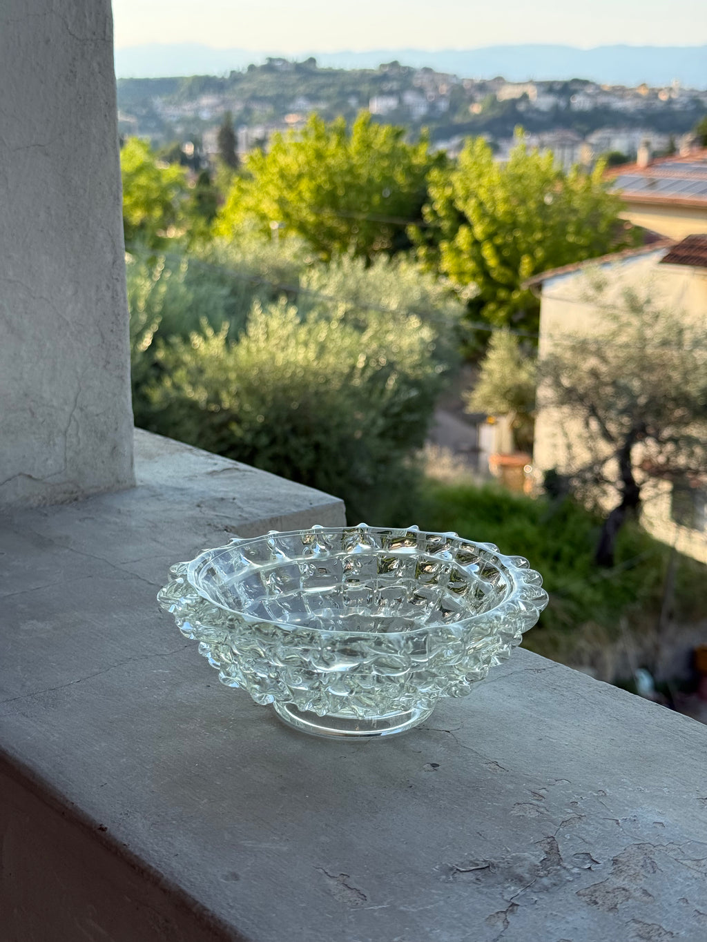 A clear textured glass bowl with a wavy, faceted rim sits on a stone balcony ledge, with a blurred outdoor garden and buildings in the background.